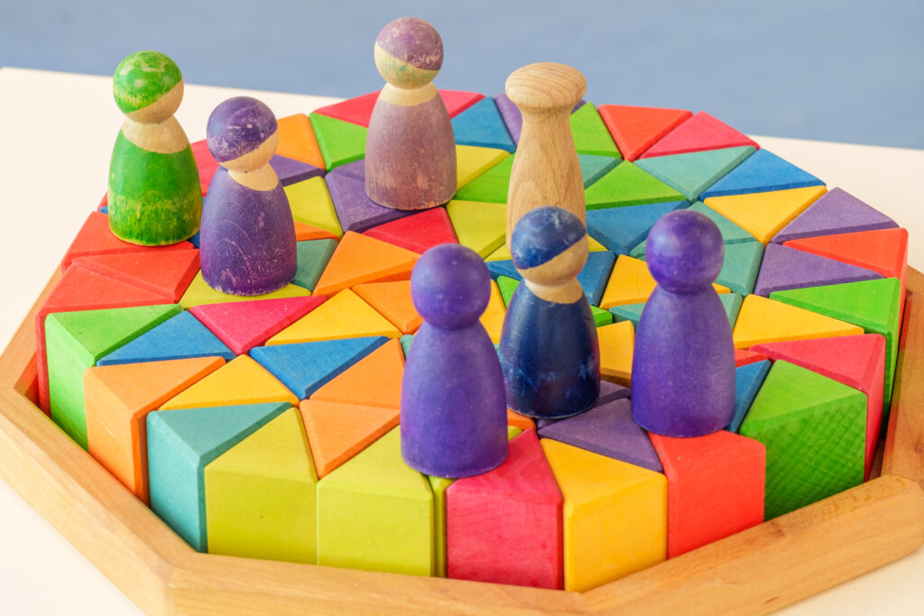 Bunte Holzbausteine in Dreieckform und verschiedenen Farben, auf denen kleine Holzfiguren stehen; pädagogisches Spielmaterial auf hellem Tisch.